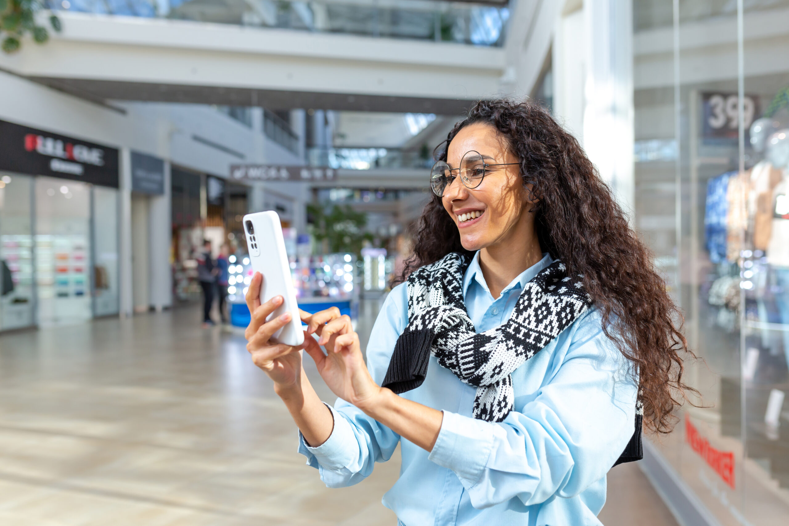 Happy woman shopping for clothes during the period of discounts and promotional offers, Hispanic woman smiling and happy browsing online discounts using smartphone, inside a modern large store